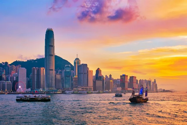Boats sailing across Victoria Harbour in front of the Hong Kong skyline at sunset.